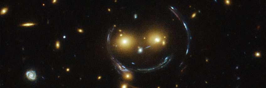 Gravitationslinseneffekt durch den Galaxienhaufen Cheshire Cat (in gelb). Dieser Haufen wirkt wie eine Linse, die das Licht einer dahinter liegenden Galaxie (in blau) in mehrere bogenförmige Bilder verzerrt, sogenannte „Einsteinringe“. Die Kombination des Galaxienhaufens im Vordergrund mit der verzerrten Hintergrundgalaxie ergibt das Bild eines lächelnden Gesichts. Bild: NASA/ESA