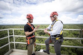 Max Planck researcher Stefan Wolff and German President Steinmeier stand on the railing of Atto's first platform. They are wearing climbing helmets and harnesses. In the background, the rainforest stretches to the horizon.