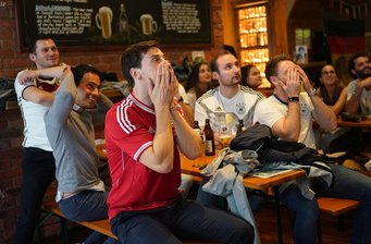 Young men and a few women, some of them wearing German national soccer team jerseys, are sitting in a pub, apparently looking at a screen outside the picture. Some of them put their hands in front of their faces or over their heads.