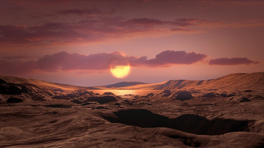 Surface of a fictitious planet. It consists of hills of rock. In the background is a lake. The sky has some clouds. Near the horizon a sun shines and bathes the picture in a reddish colour.