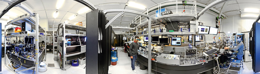 Panoramic image of a laboratory showing three experimental tables, each several square metres in size, each with numerous optical devices such as lasers, mirrors, etcetera, as well as computer screens. At one table a man is adjusting an optical device, at the same table another man is sitting in front of a computer. Both people are wearing protective goggles against the laser light.

