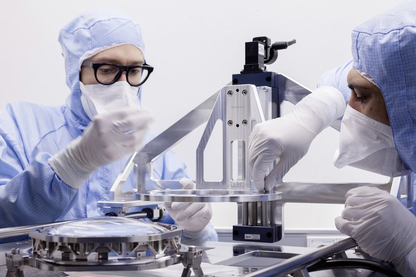 Close-up of two people in surgical clothing and FFP masks working on a shiny, bright metal apparatus. On the left of the picture is an optical lens, about the size of the palm of the hand, set in a metal frame, on the right is the metal frame on which the lens system is to be placed. 
