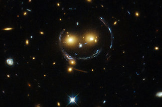 Gravitationslinseneffekt durch den Galaxienhaufen Cheshire Cat (in gelb). Dieser Haufen wirkt wie eine Linse, die das Licht einer dahinter liegenden Galaxie (in blau) in mehrere bogenförmige Bilder verzerrt, sogenannte „Einsteinringe“. Die Kombination des Galaxienhaufens im Vordergrund mit der verzerrten Hintergrundgalaxie ergibt das Bild eines lächelnden Gesichts. Bild: NASA/ESA