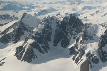 Aerial view of a mountain massif with a basin in the foreground, glaciers stretching to the horizon behind the mountain.