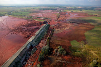 A rectangular red mud landfill can be seen in the left half of the picture. Approximately in the centre of the picture you can see that the dam of the landfill has broken at one corner, flooding the surrounding landscape with red mud over an area about the size of the landfill itself.